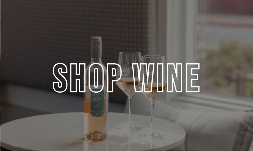 Shop Wine text overlaid an image of a wine bottle on a table with two glasses, near a bright window
